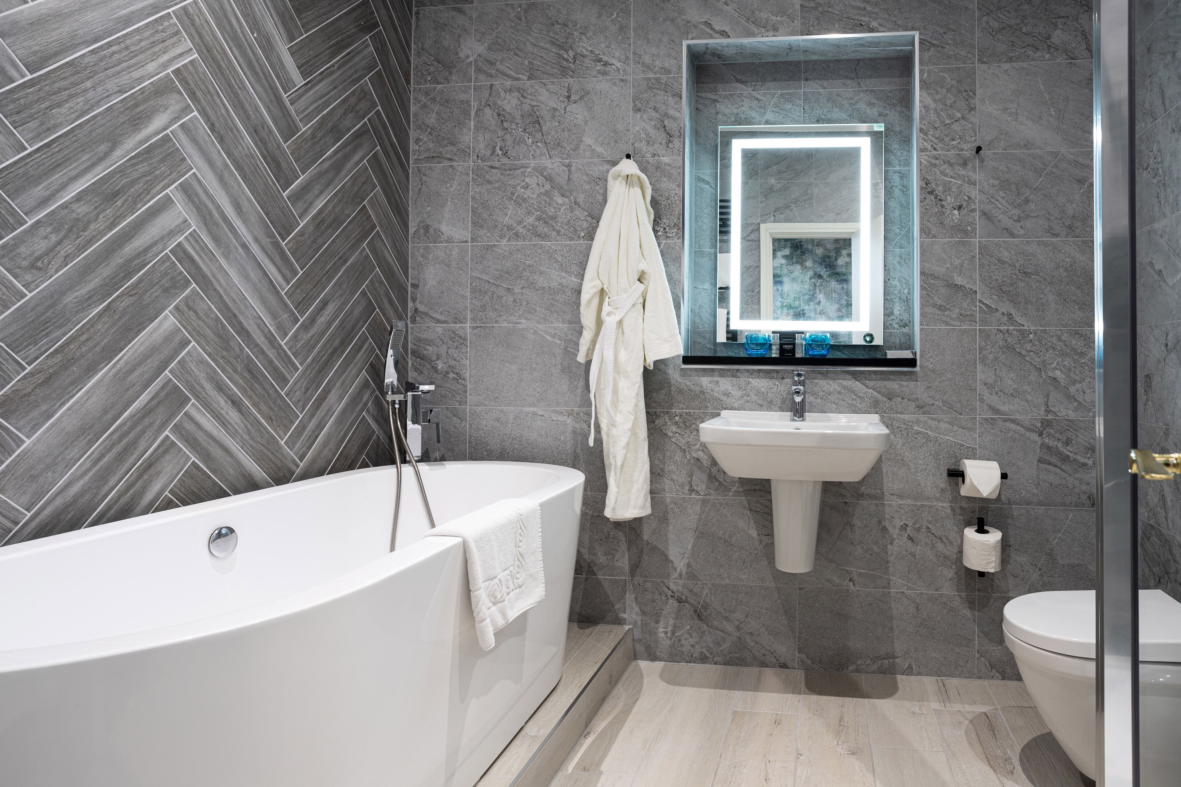 A modern bathroom with a herringbone-patterned wall, a freestanding bathtub, a pedestal sink, and a toilet.