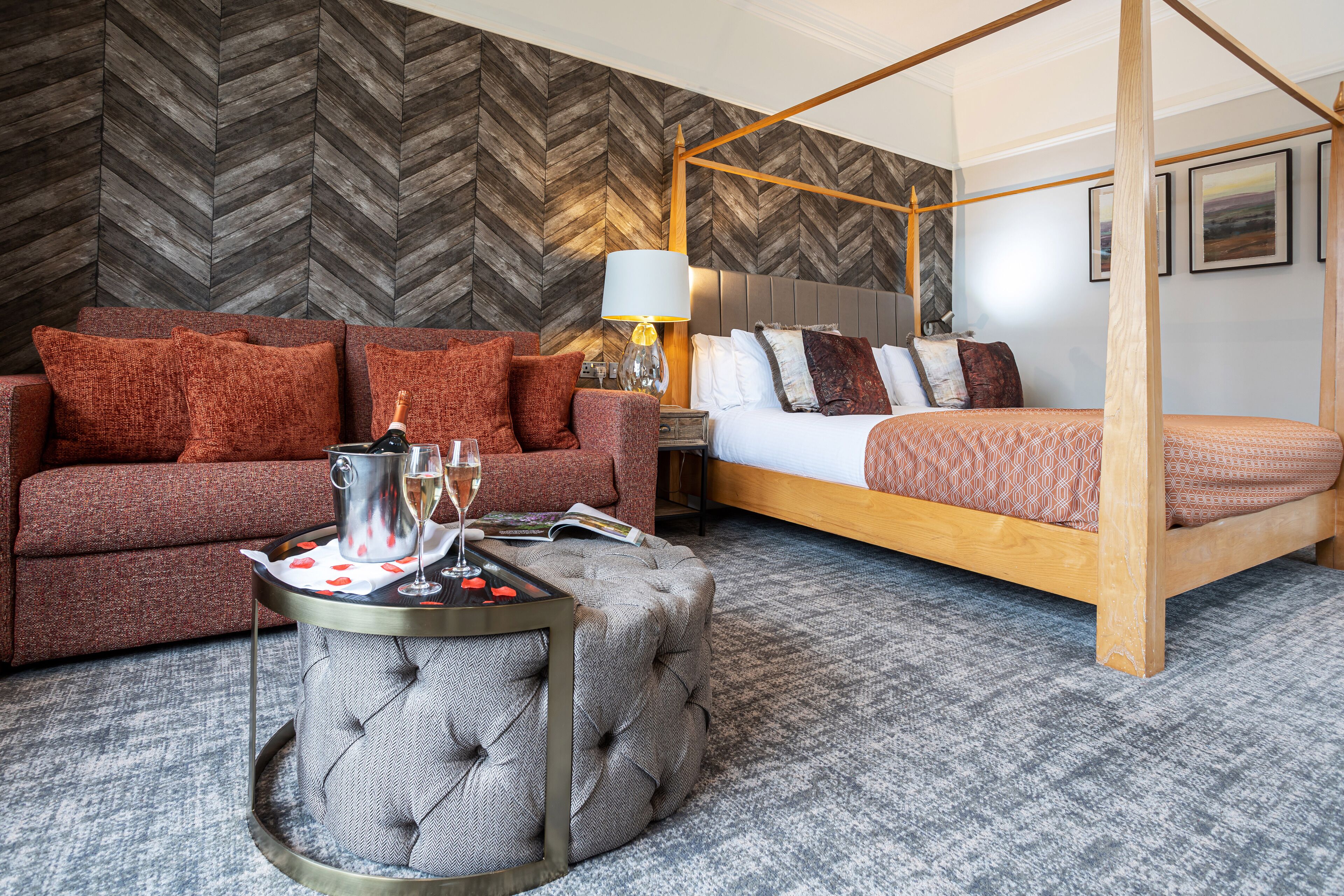 A modern hotel room with a herringbone-patterned wall, a four-poster bed, a tufted ottoman, and a sofa with red cushions.