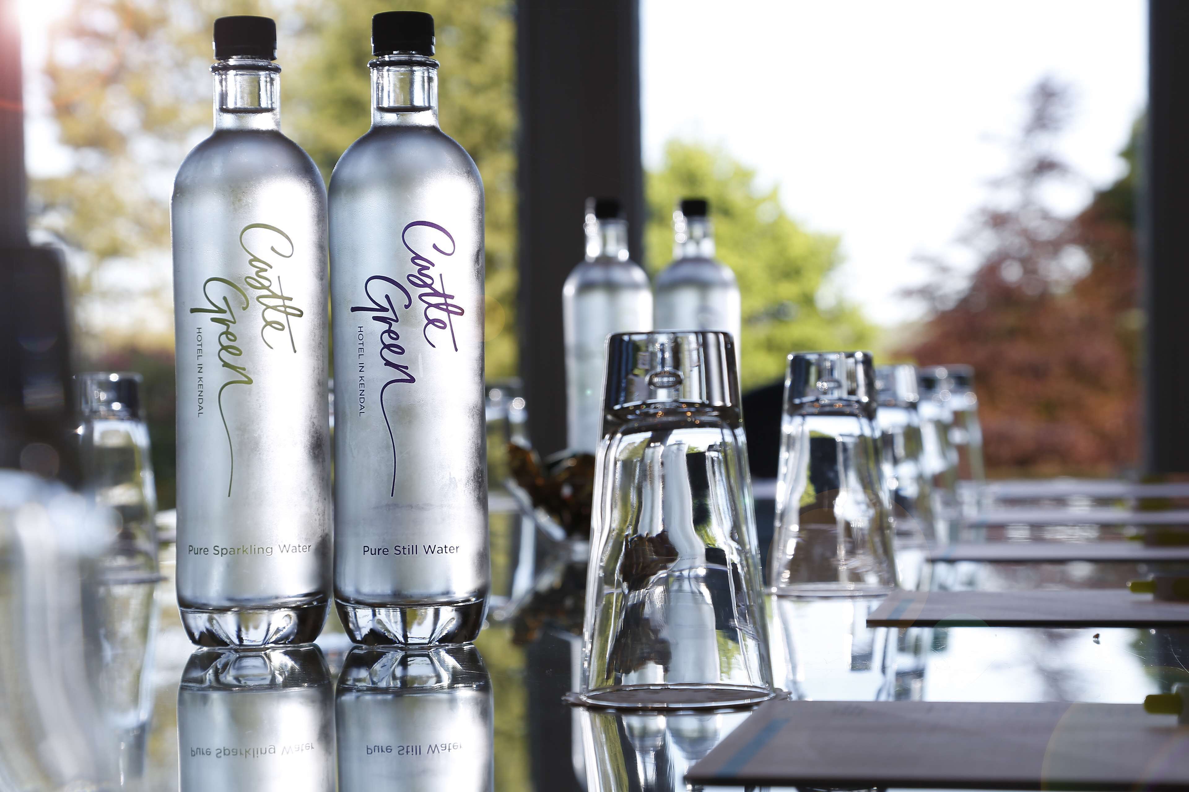 Dos botellas de agua Cotte Green, una etiquetada como "Agua Con Gas Pura" y otra como "Agua Mineral Sabor Original". Se observan vasos de vidrio transparentes sobre una superficie reflectante al fondo.