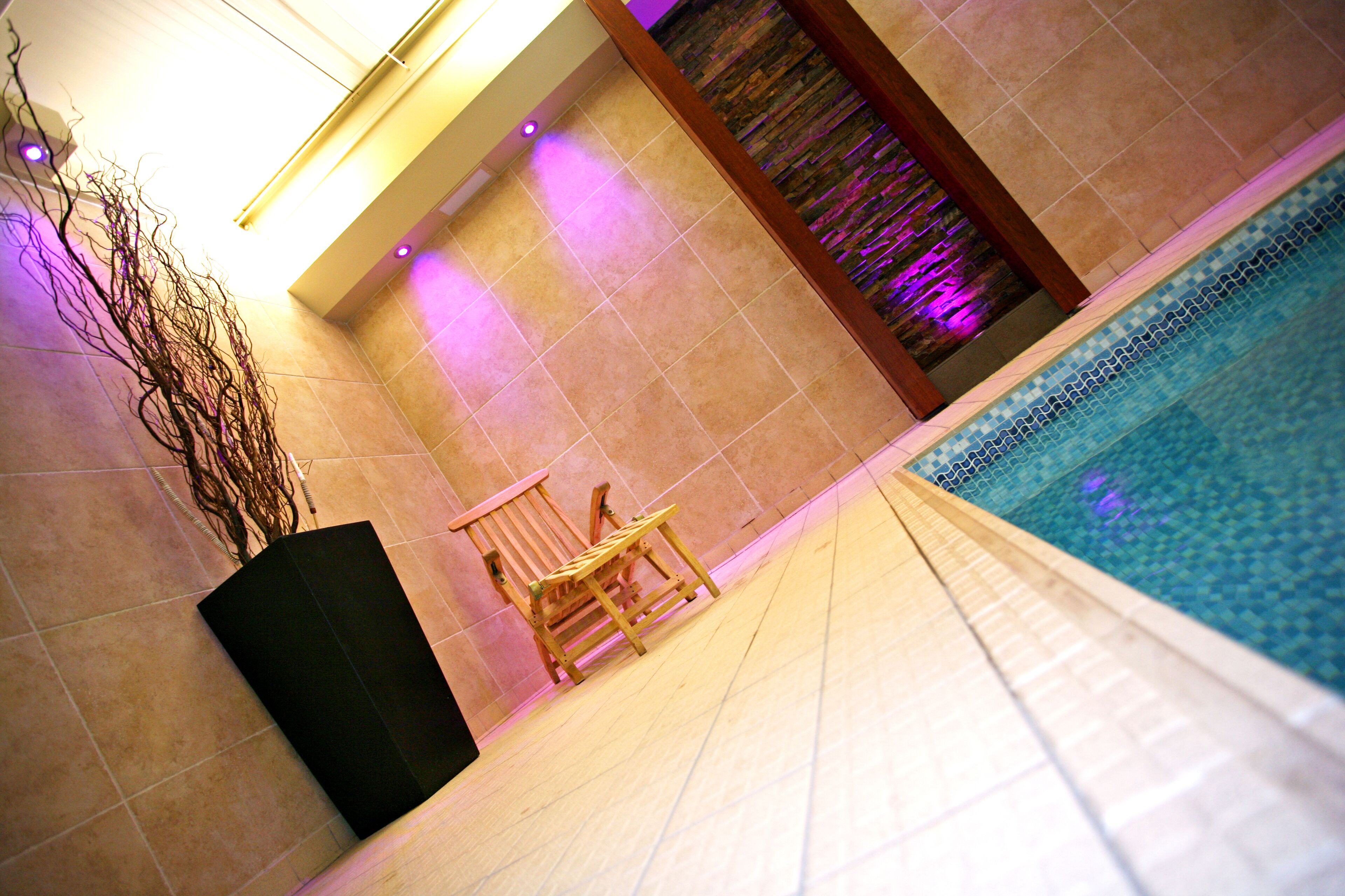 A swimming pool area with tiled flooring, a wooden chair, and a decorative wall with a water feature.