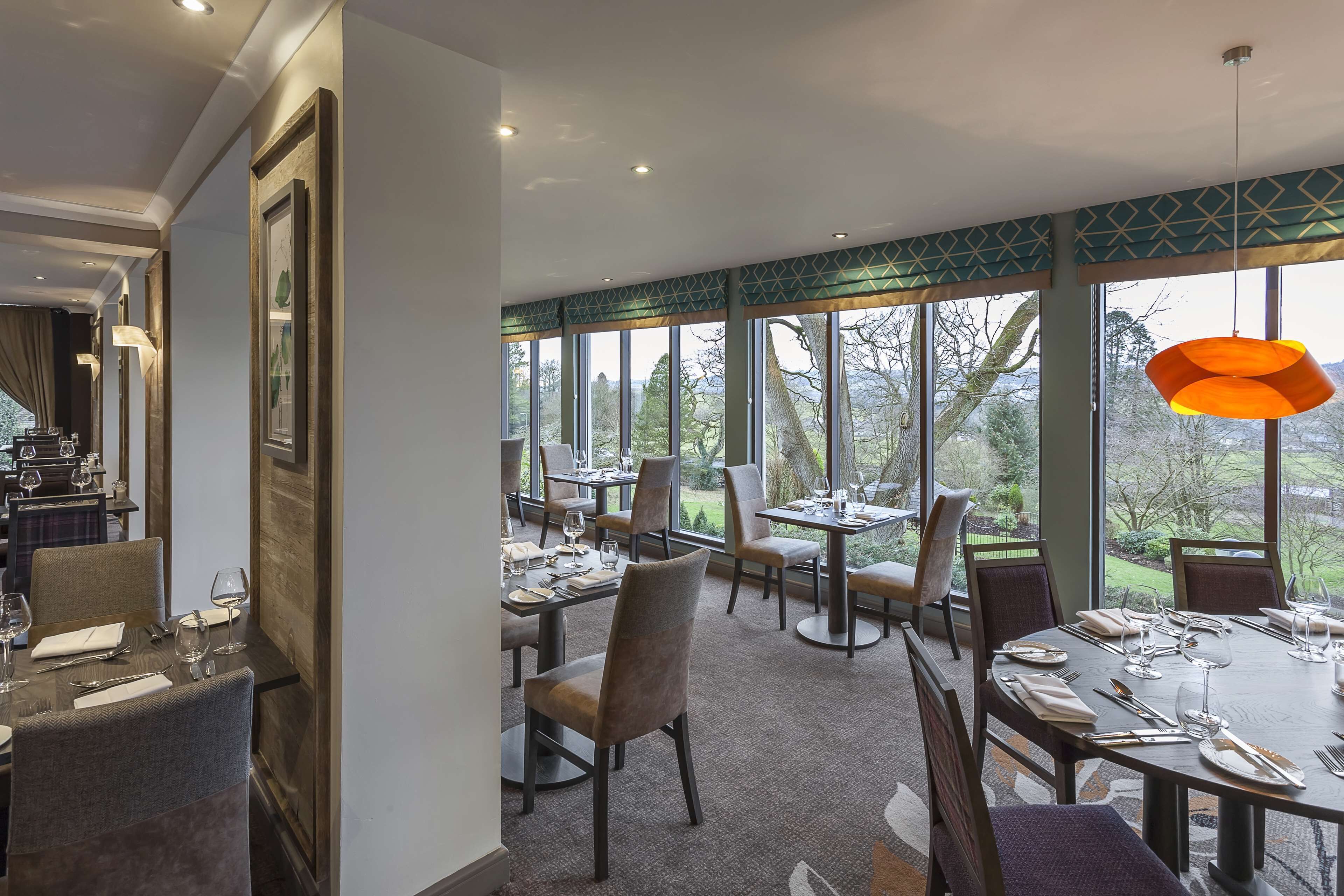 Un espace repas avec des tables dressées pour le repas, de grandes fenêtres offrant une vue sur l’extérieur et un luminaire suspendu.