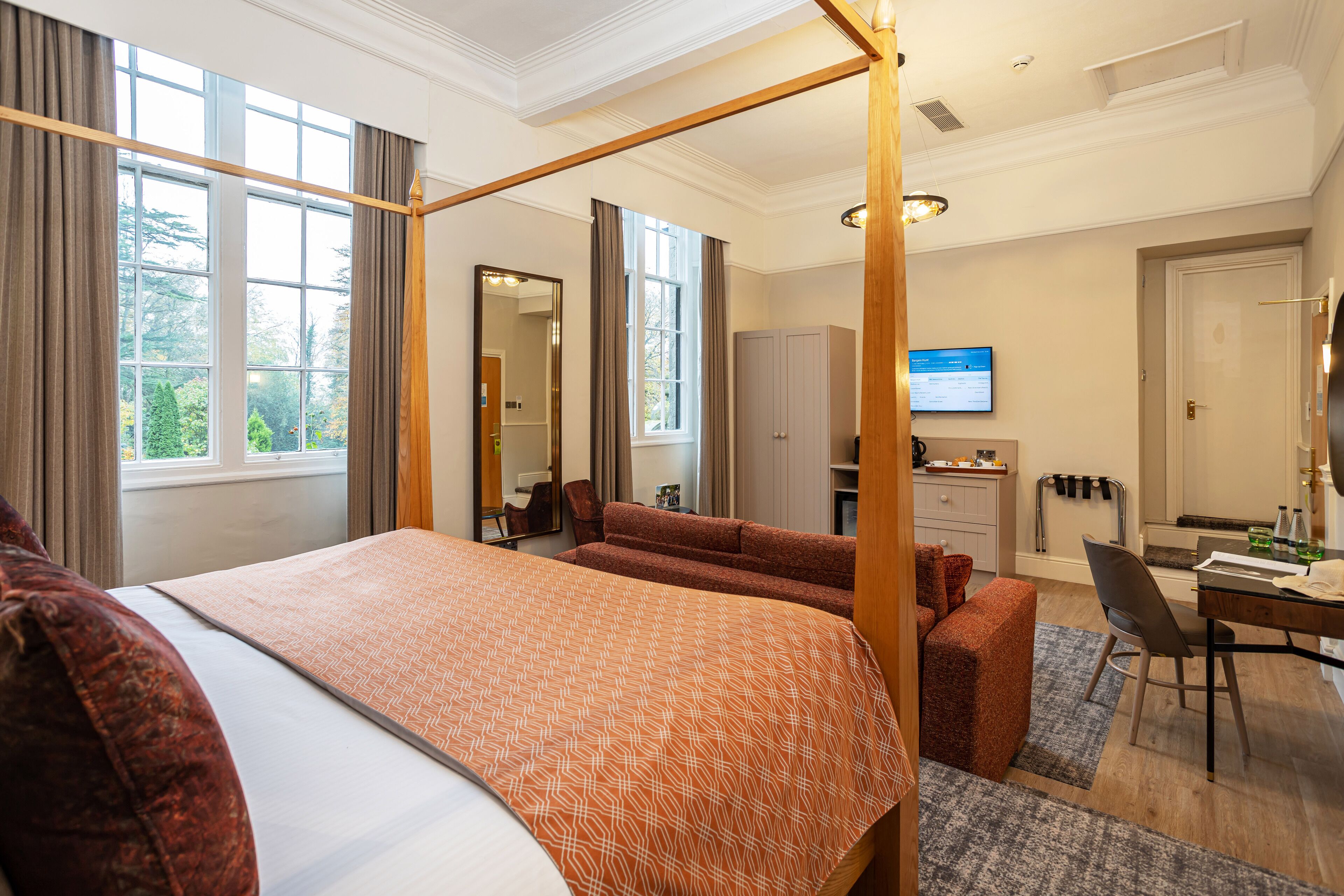 A hotel room with a four-poster bed, a desk, a chair, a sofa, and a television.