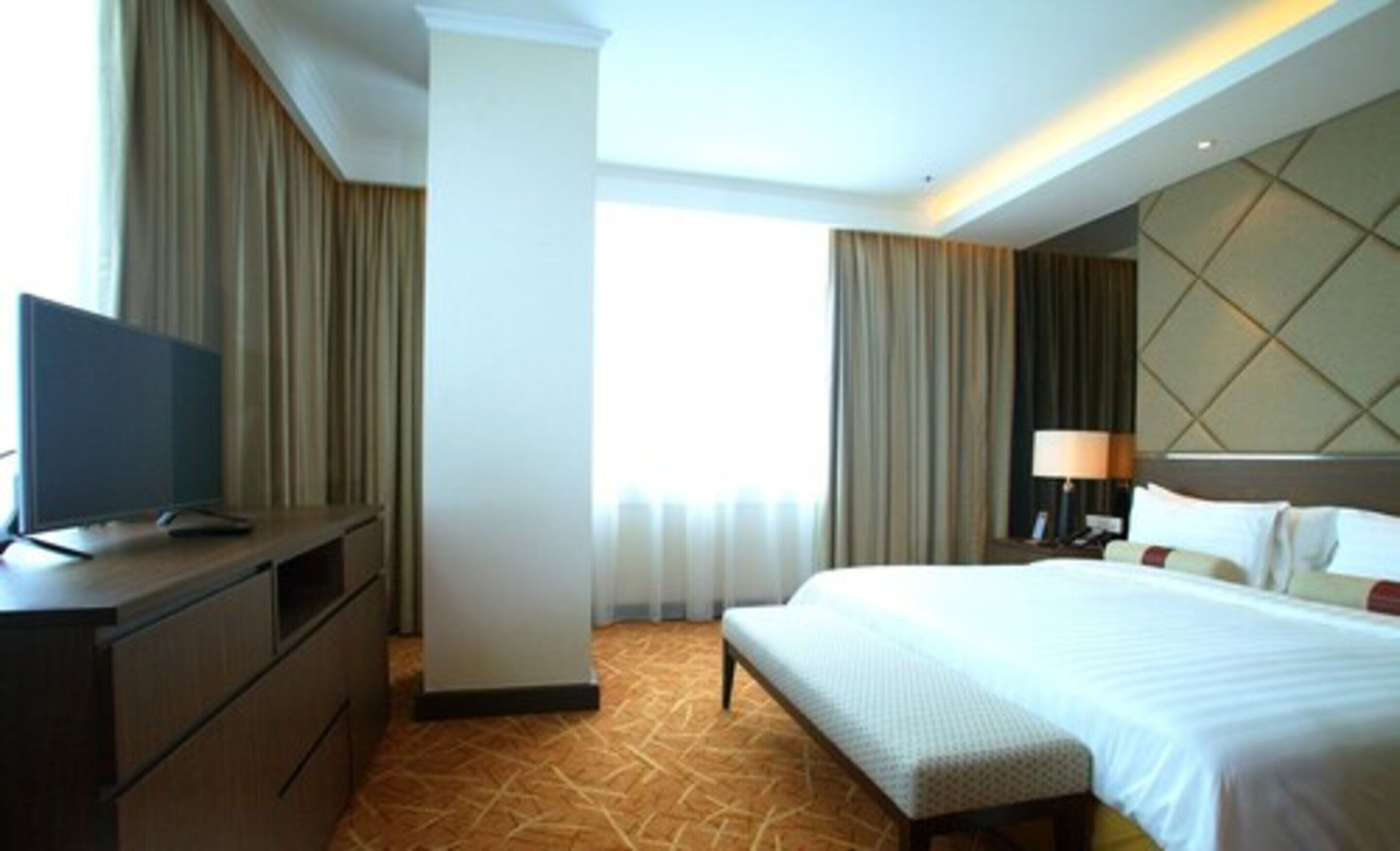 A hotel room with a bed, a TV on a desk, a lamp, and curtains.