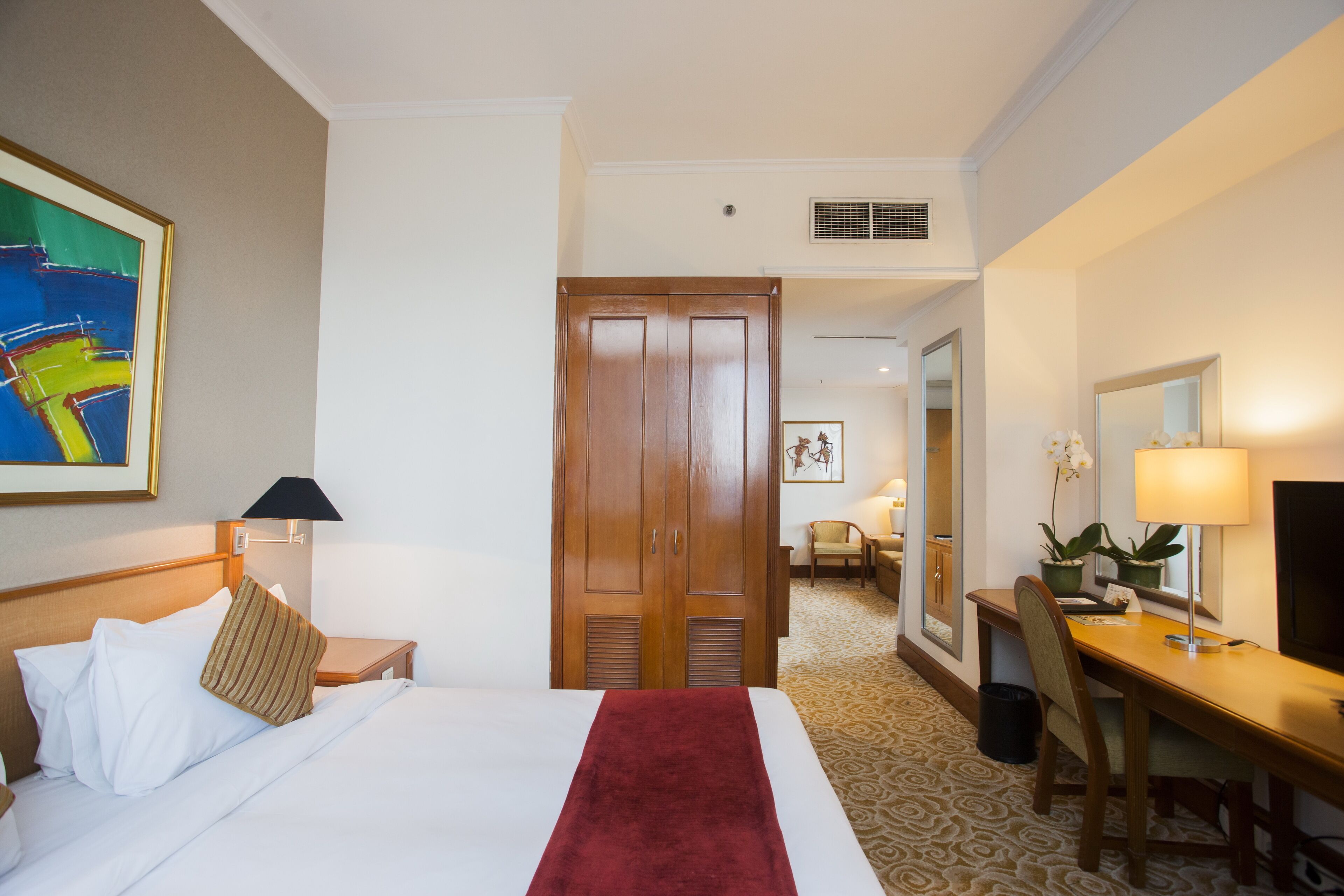 Une chambre d’hôtel comprenant un lit, un bureau, une chaise, une lampe de bureau, une plante en pot et un miroir.