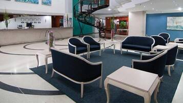 A hotel lobby with a reception desk, seating area with chairs and tables, a staircase, and artwork on the walls.