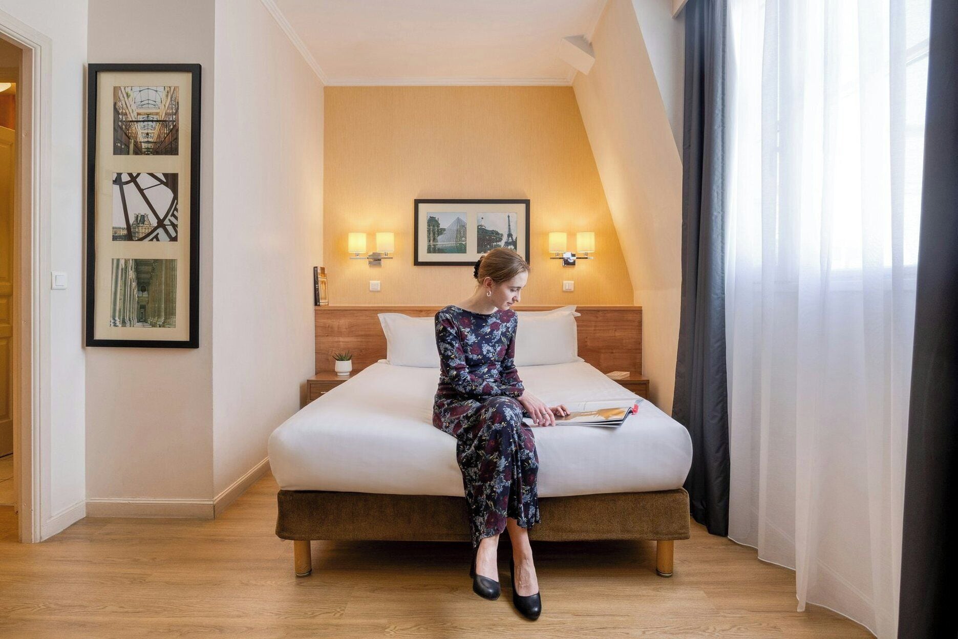 Une chambre d’hôtel avec un lit, une table de chevet, une lampe fixée au mur et des tableaux encadrés.