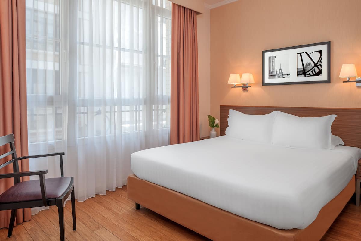 Habitación de hotel con una cama grande, una silla, un ventanal con cortinas translúcidas y un cuadro enmarcado en la pared.