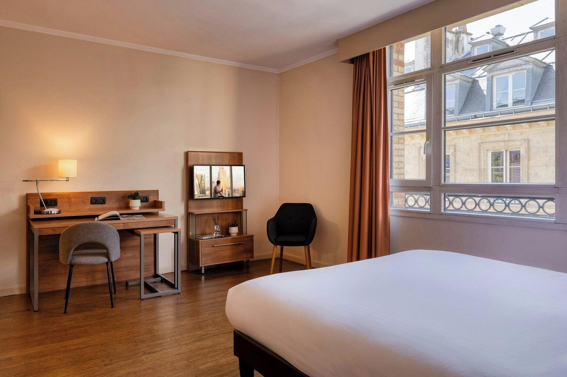 A hotel room with a bed, desk, chair, and a large window with a view of buildings.