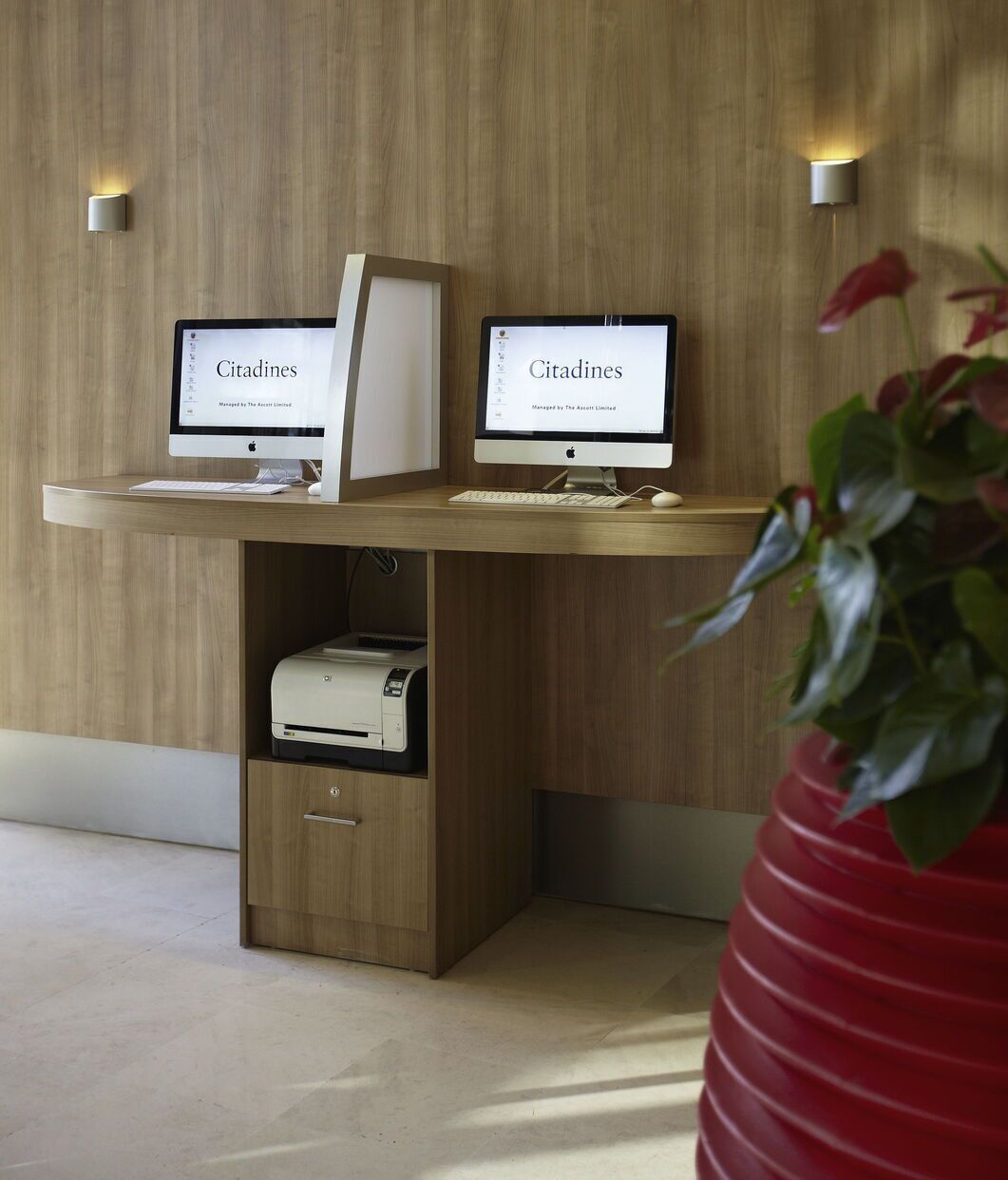 Two Apple iMac computers on a wooden desk with a printer drawer below, against a wooden wall with two wall-mounted lights.