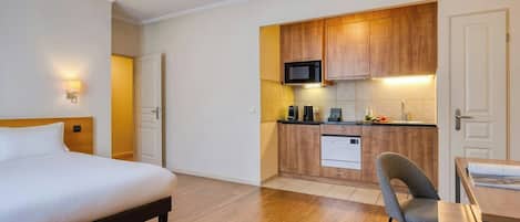 A hotel room with a bed, a small kitchenette, a dining area with a table and chair, and a door.