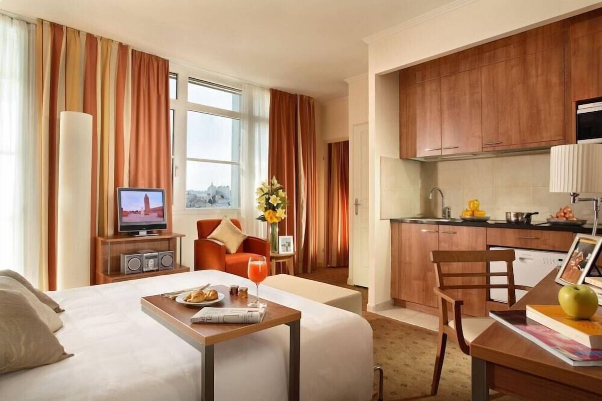 Habitación de hotel con cama, televisión, mesa de desayuno, cocina y vista panorámica de la ciudad.