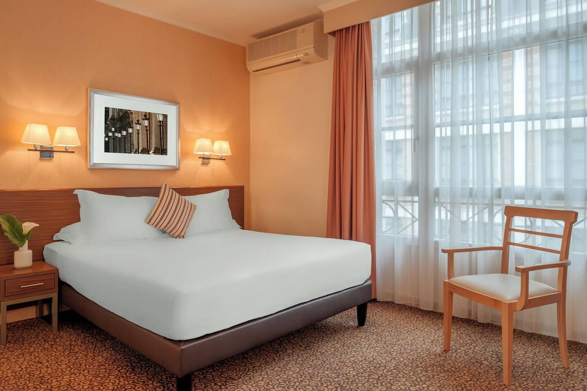Una habitación de hotel con una cama grande, dos lámparas de noche, un cuadro enmarcado sobre la cama, una ventana con cortinas traslúcidas y una silla de madera.