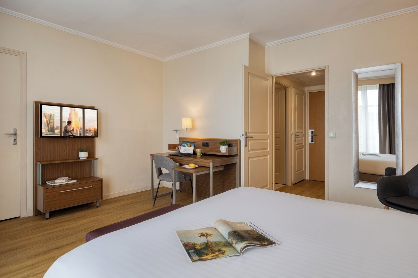 A hotel room with a bed, a desk, a chair, a small cabinet, a mirror, and a door leading to another room.