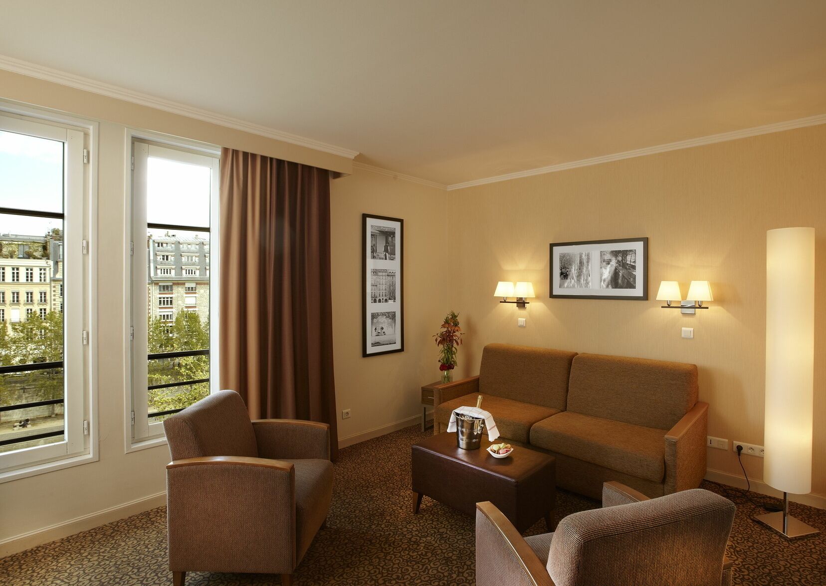 A hotel room with a sofa, two armchairs, a coffee table, and a large window with curtains. There are framed pictures on the wall and a lamp on the side table.