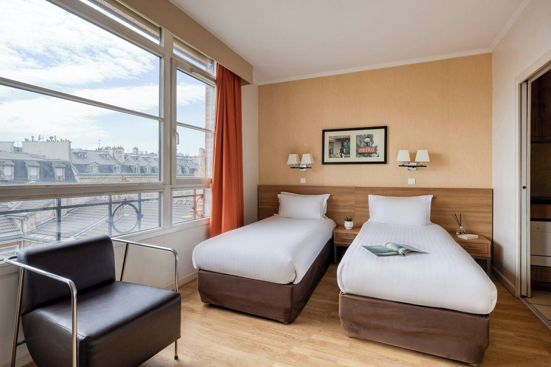 A hotel room with two beds, a chair, a window with a view of buildings, and wall-mounted lamps.