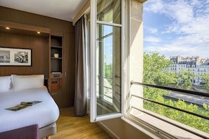 Habitación de hotel con una ventana grande, una cama, una estantería de madera y un cuadro en la pared.
