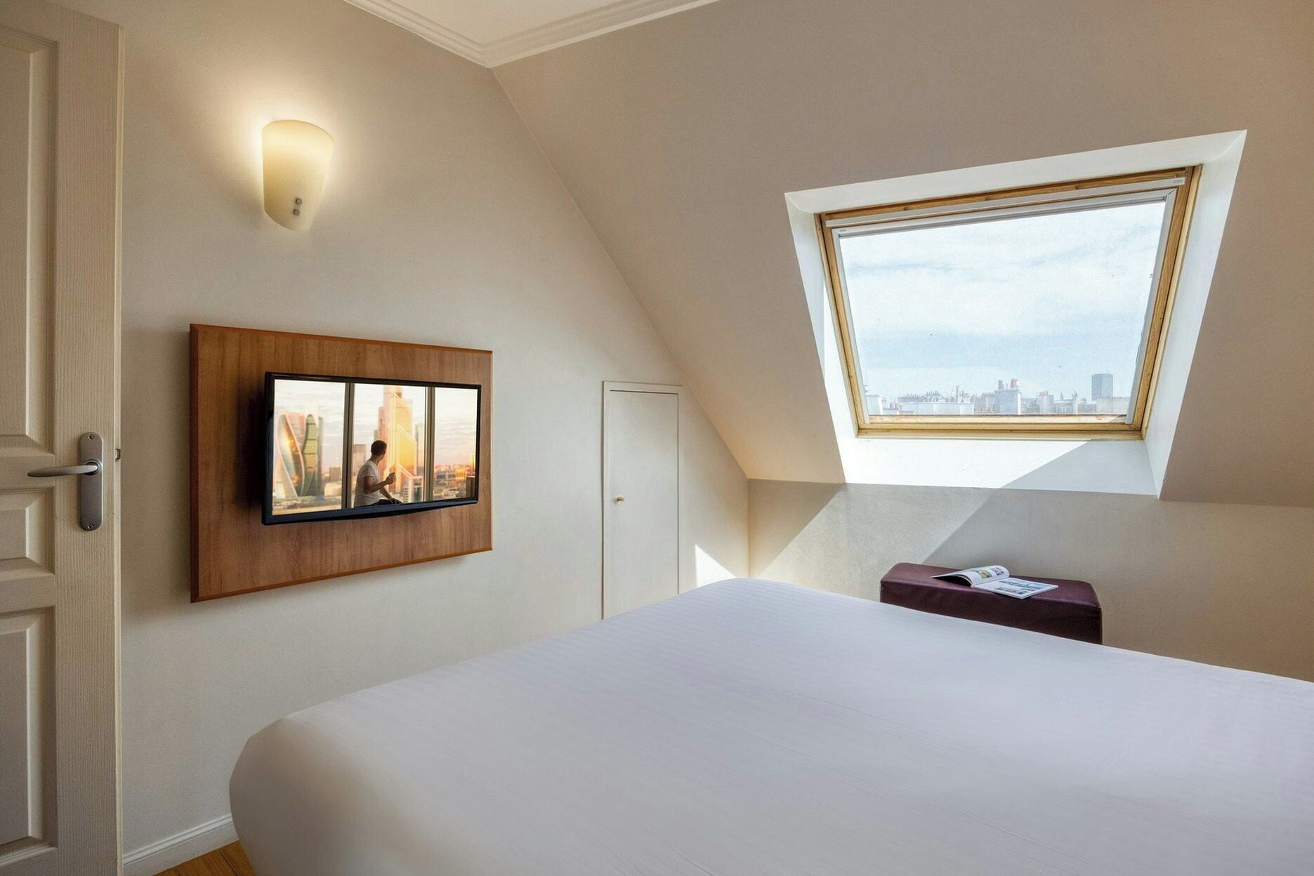 A bedroom with a large bed, a wall-mounted TV, a skylight, and a city view.