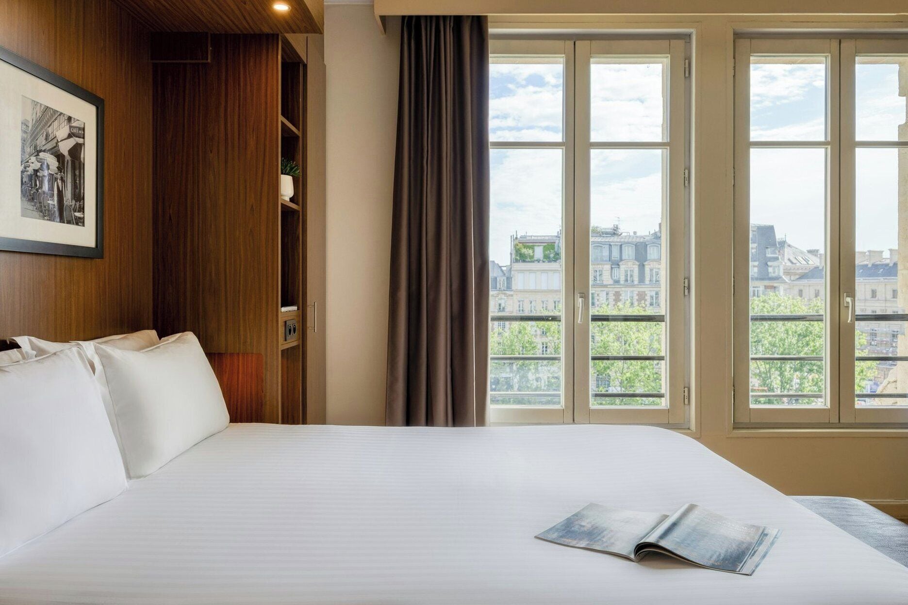 Hotelkamer met een groot bed, een raam met gordijnen, een lijst met een foto aan de muur, en uitzicht op gebouwen.