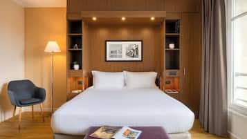 Une chambre d’hôtel avec un grand lit, une table de chevet, une chaise, une lampe sur pied et des étagères intégrées agrémentées d’un tableau encadré.