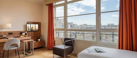 A hotel room with a desk, two beds, a chair, a TV, and a large window with curtains.