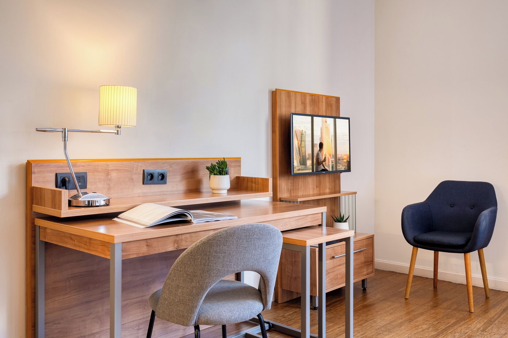 Een moderne hotelkamer met een houten bureau, een stoel, een lamp, een flatscreen televisie en een plant in pot.