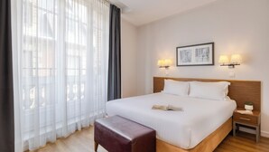 Une chambre d’hôtel avec un grand lit, une tête de lit en bois, deux lampes de chevet, une table de chevet, une fenêtre avec des rideaux légers et un tableau encadré au mur.
