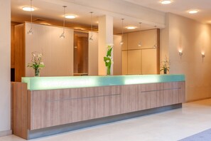 Ein moderner Empfangstresen mit heller Holzoptik und einer Glasplatte. Auf dem Tresen stehen zwei Blumentöpfe, und der Raum zeichnet sich durch ein aufgeräumtes, minimalistisches Design mit Holzpaneelen und eingelassener Beleuchtung aus.