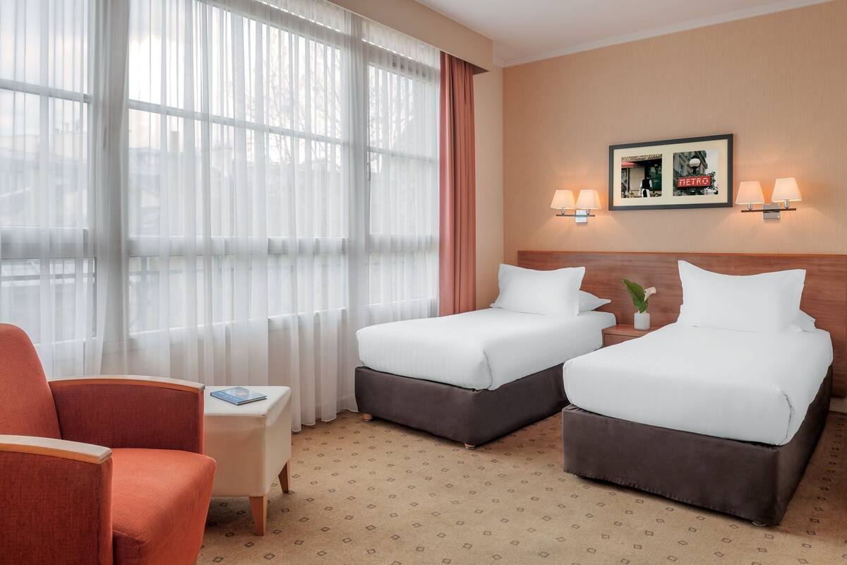 Habitación de hotel con dos camas, una silla, una mesita y un cuadro colgado en la pared.