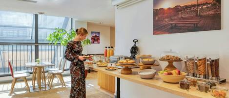 Habitación de hotel con bufé de desayuno, un ventanal con vistas y un cuadro de un paisaje urbano.