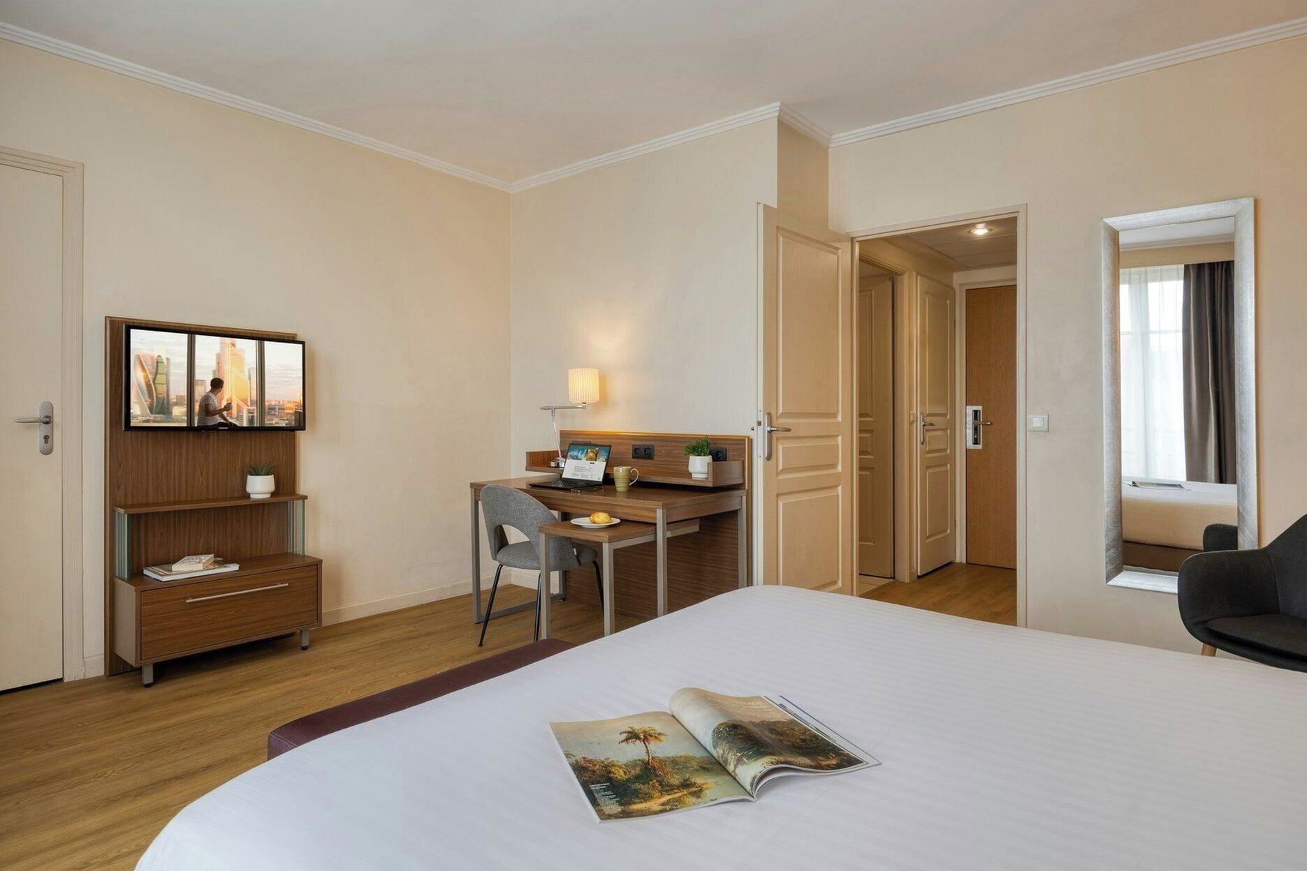A hotel room with a bed, a desk, a chair, a bookshelf, and a window with curtains.