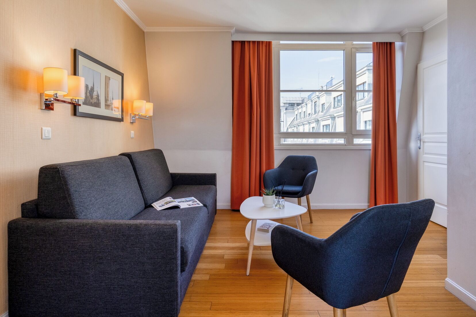 Una habitación moderna con un sofá gris, una pequeña mesa redonda, una silla azul y una ventana con cortinas.