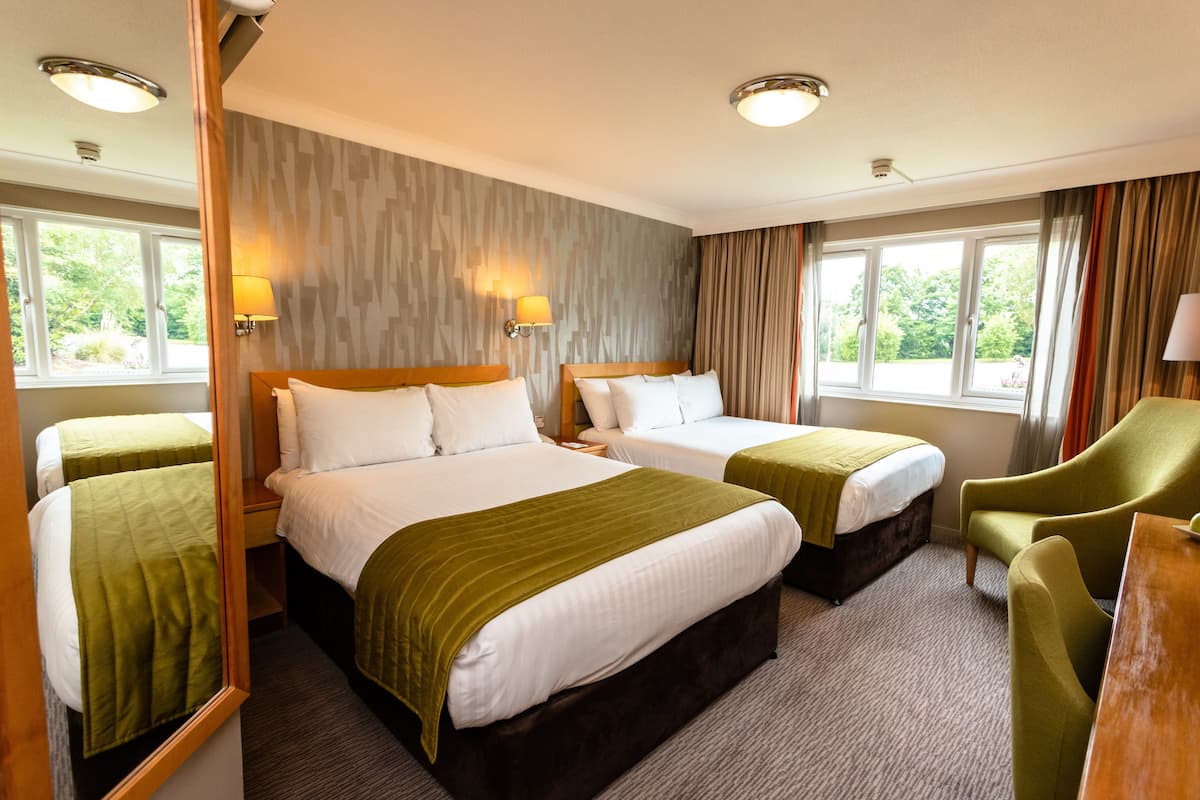 Habitación de hotel con dos camas individuales, una cama grande y un sillón verde. Hay dos mesitas de noche y un escritorio con una silla.