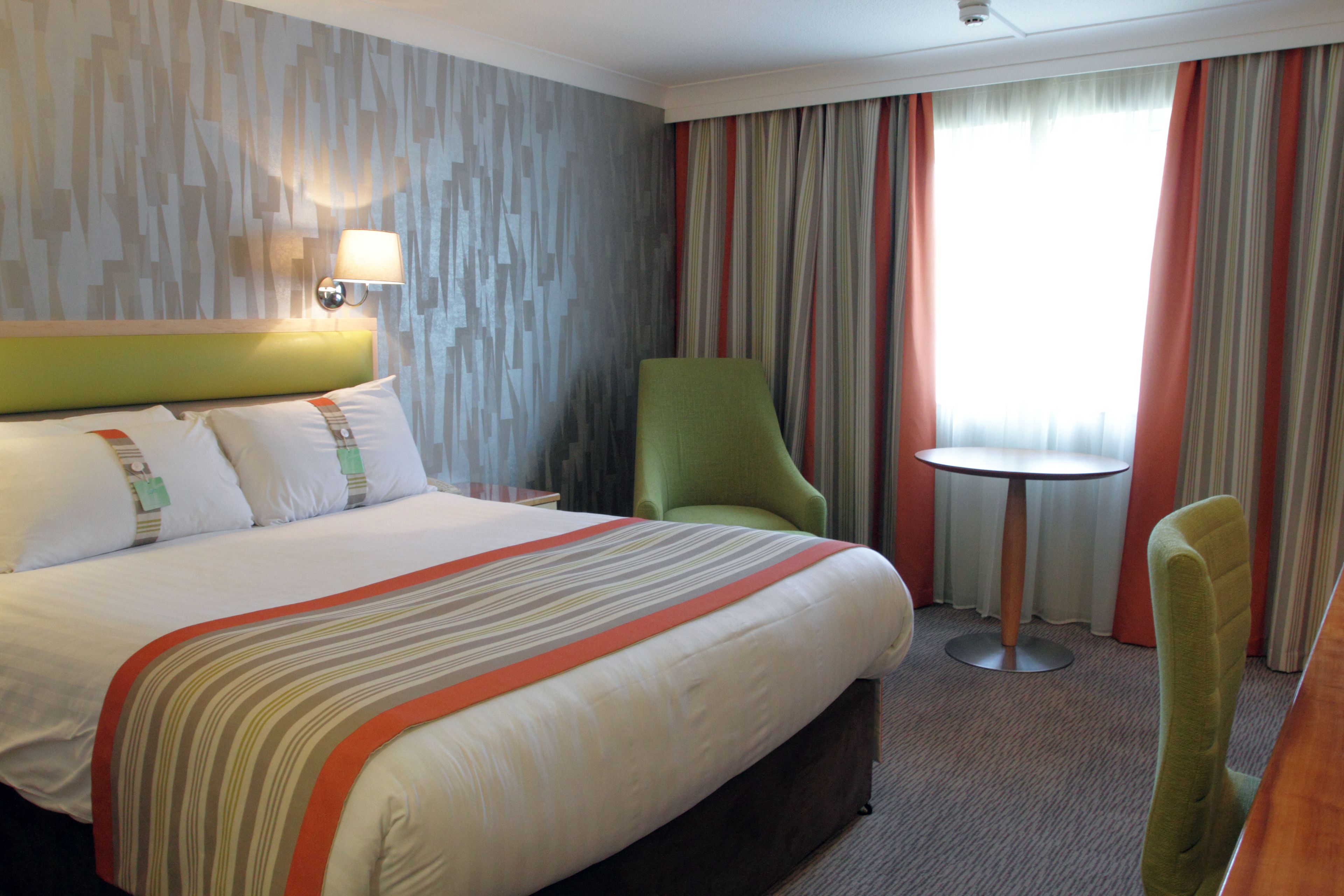A hotel room with a large bed, a green armchair, a small round table, and a view of the outdoors through curtains.