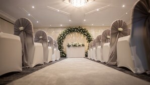 A carpeted aisle with rows of chairs on either side, a floral arch at the end, and a chandelier above. - Holiday Inn A55 Chester West by IHG (Deeside)