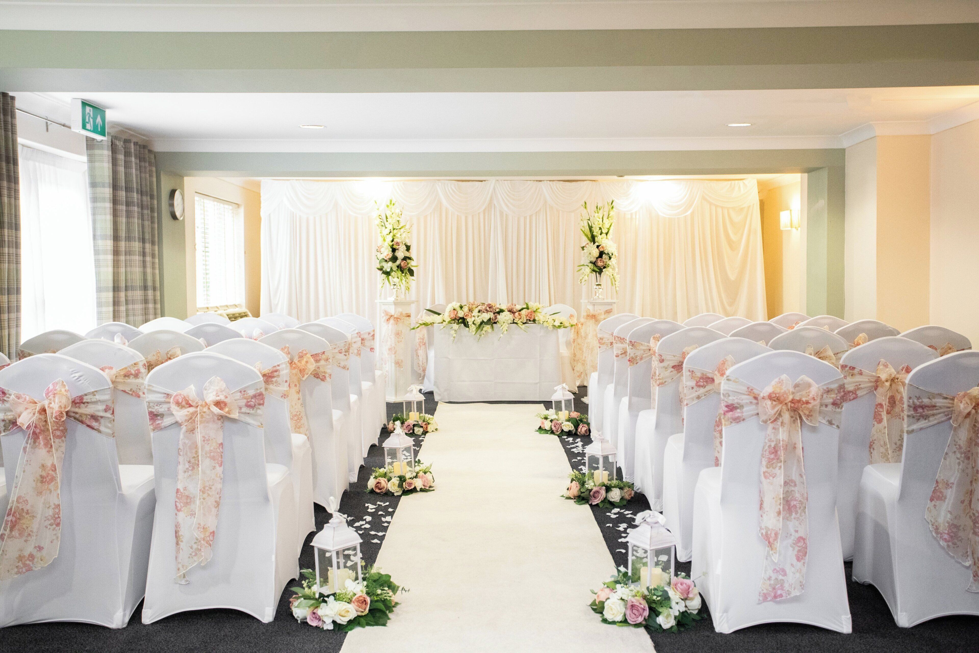 A hotel conference room with rows of white chairs, floral chair covers, and a black carpet aisle.
