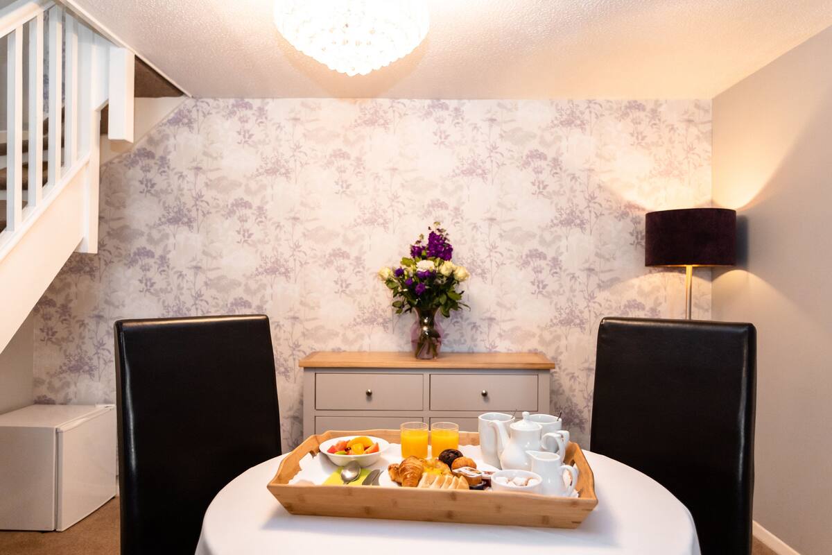 Una bandeja de desayuno con croissants, fruta y jugo sobre una mesa en una habitación con papel tapiz floral, un jarrón de flores y una lámpara.
