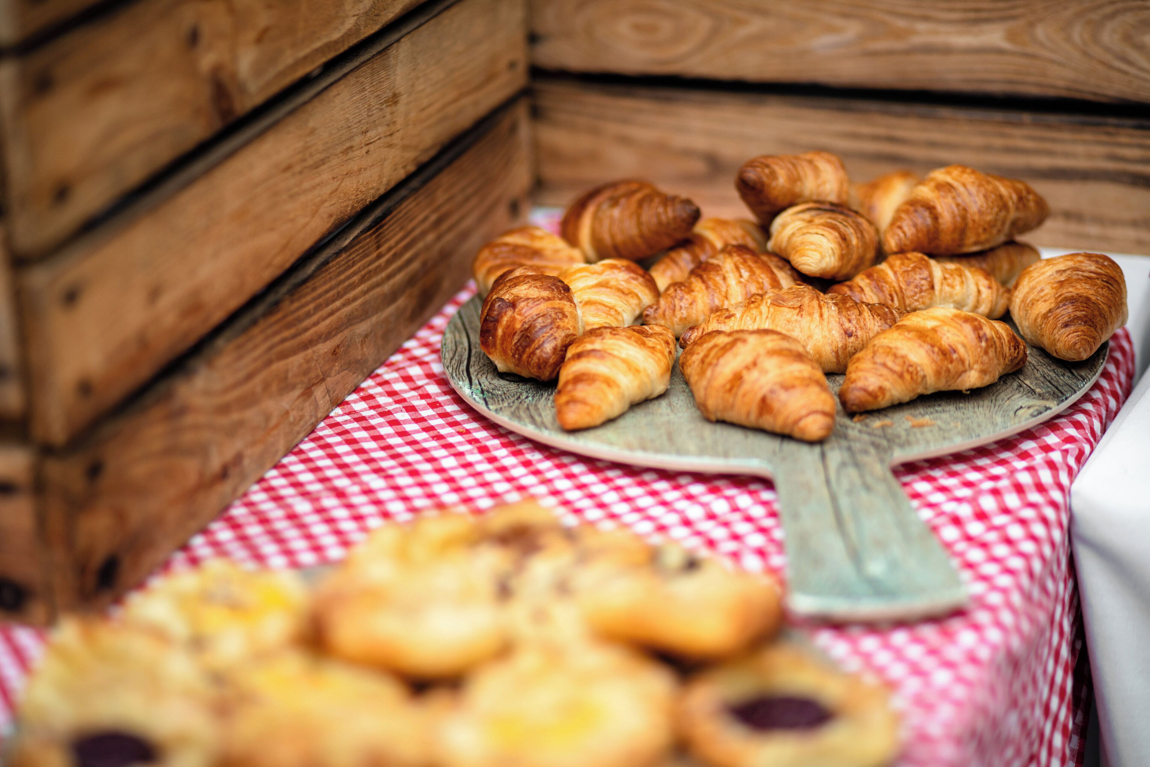 A wooden crate filled with pastries, a plate of croissants on a checkered tablecloth, and a blurred section of baked goods.