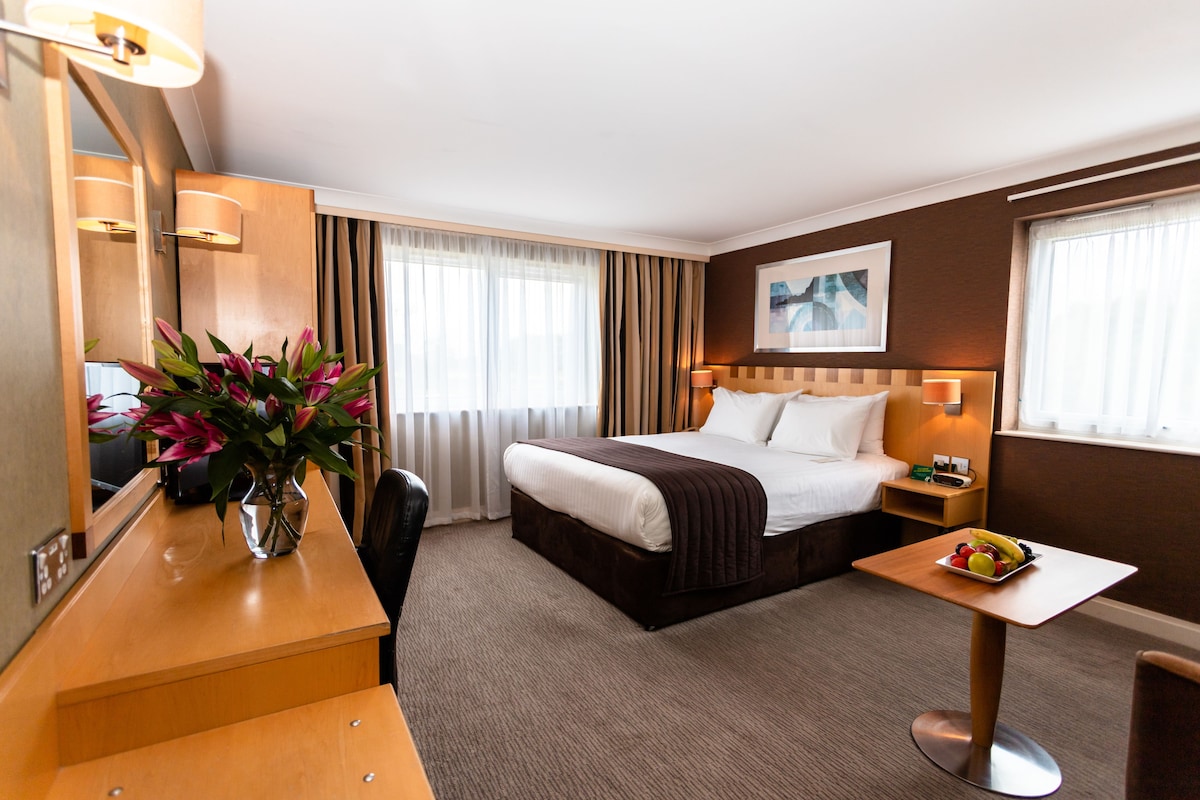 A hotel room with a bed, bedside table, desk, chair, and a vase of flowers.