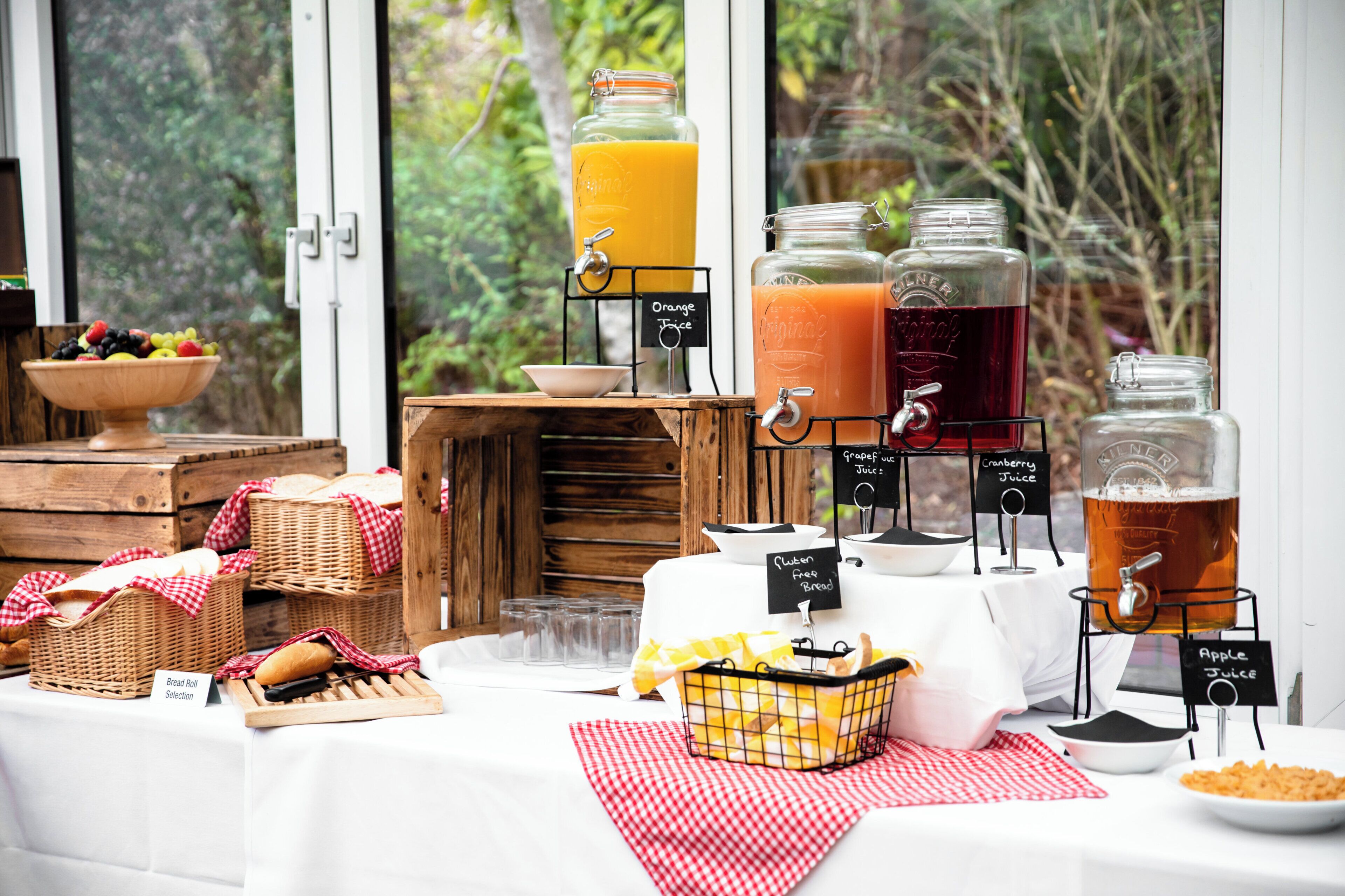 A buffet table with various food and drink options, including a selection of bread, a fruit bowl, and multiple beverage dispensers.