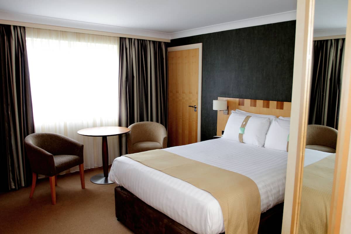 Una habitación de hotel con una cama grande, dos sillones, una mesita redonda y una puerta.