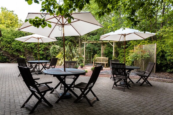 An outdoor patio with tables and chairs, surrounded by greenery and a wooden pergola.