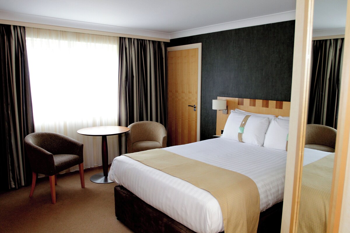 Une chambre d’hôtel avec un lit, deux fauteuils, une petite table ronde et une porte.