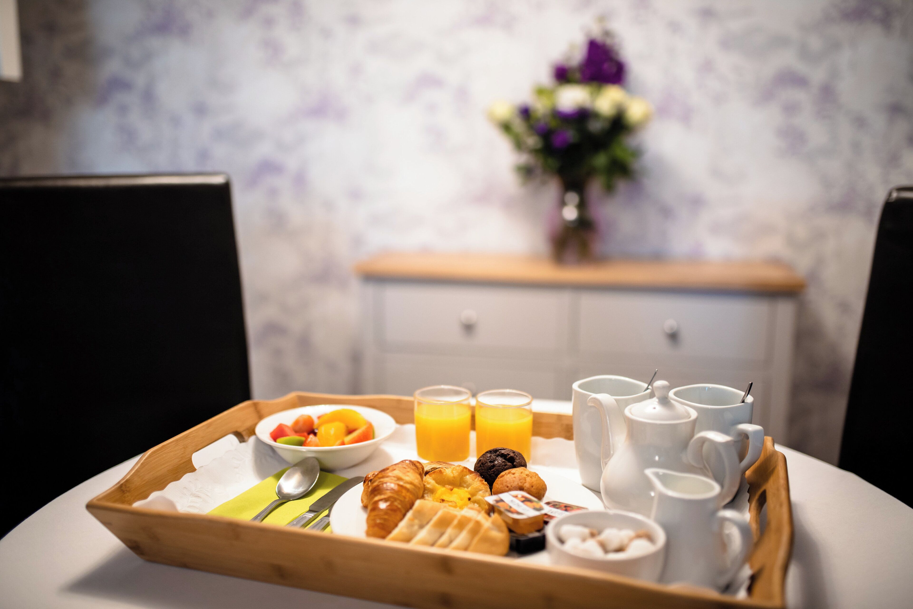 Un plateau de petit-déjeuner avec des croissants, des fruits et du jus, posé sur une table agrémentée de tasses blanches et d’un vase de fleurs en arrière-plan.