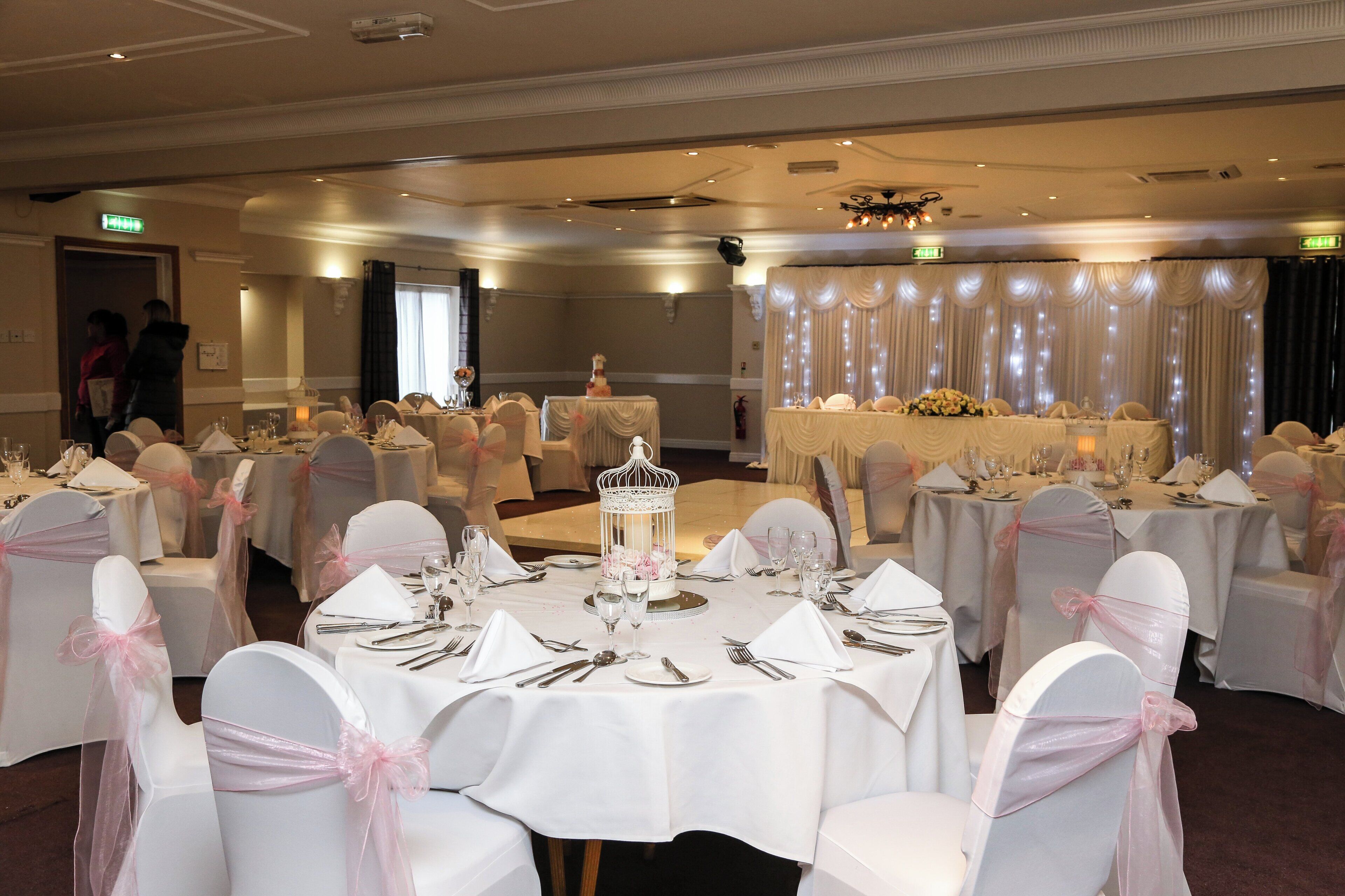 Un salón de banquetes con mesas redondas dispuestas para un evento formal, con manteles blancos y sillas adornadas con cintas rosas.