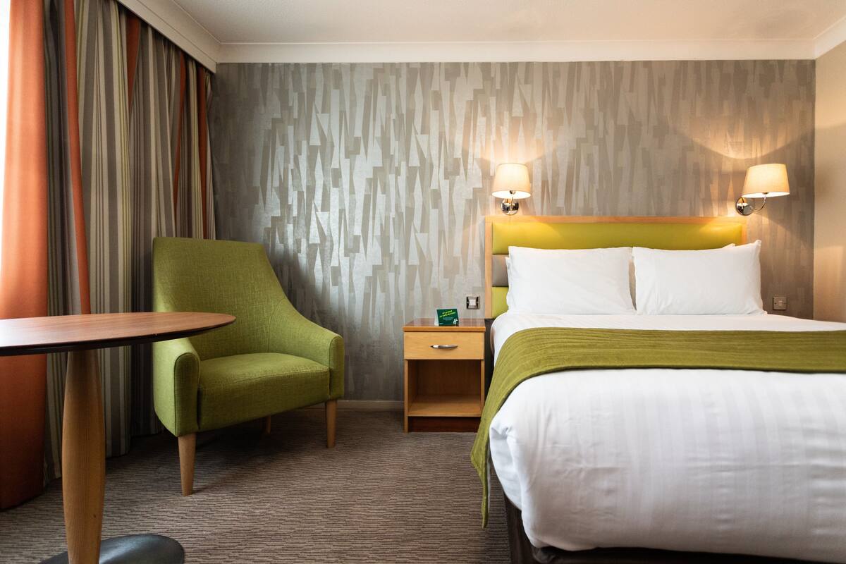 Una habitación de hotel con una cama, un sillón verde, una mesita de noche de madera y una mesa.