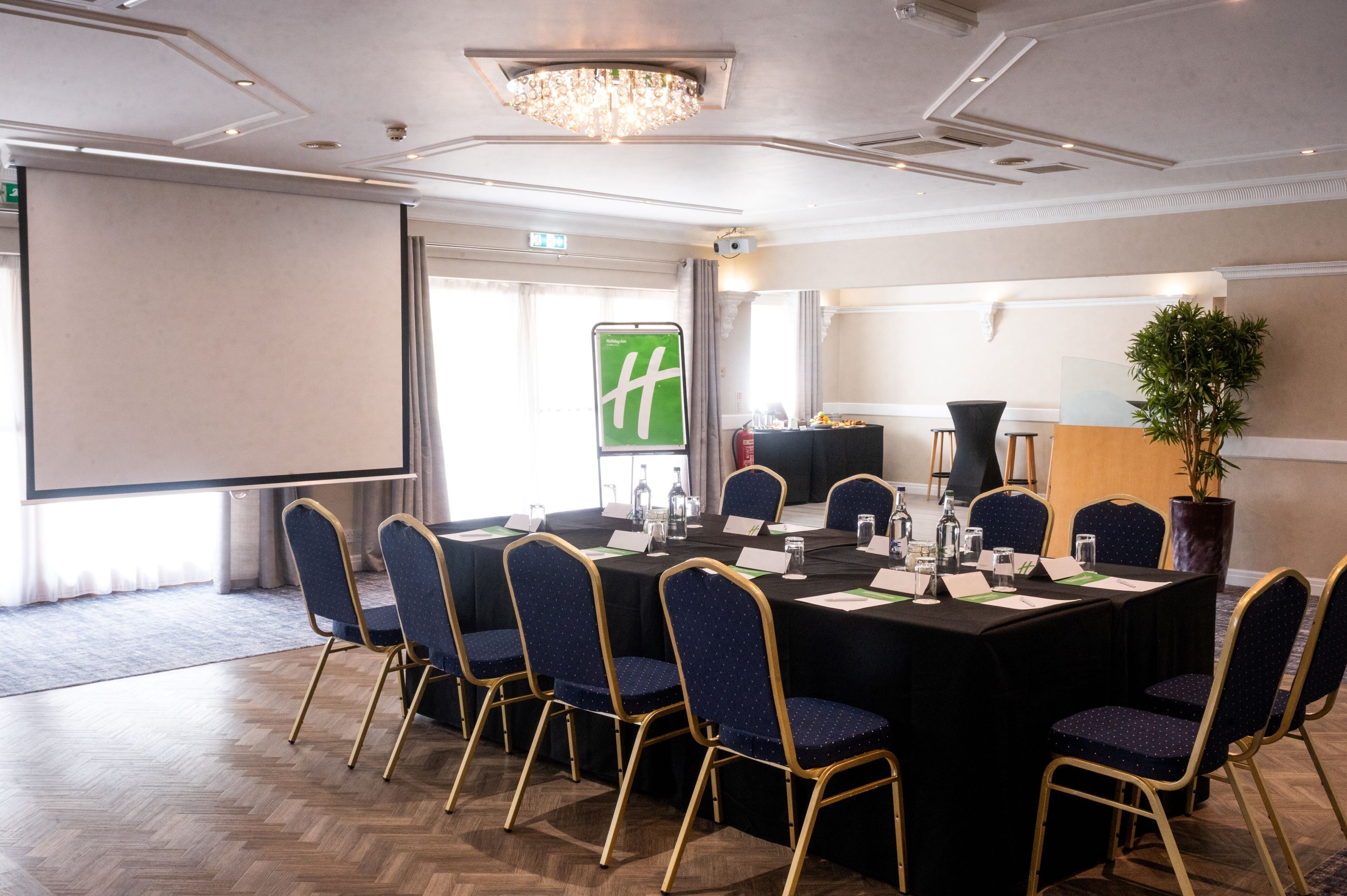 A conference room with a large table, chairs, a projector screen, and a plant.