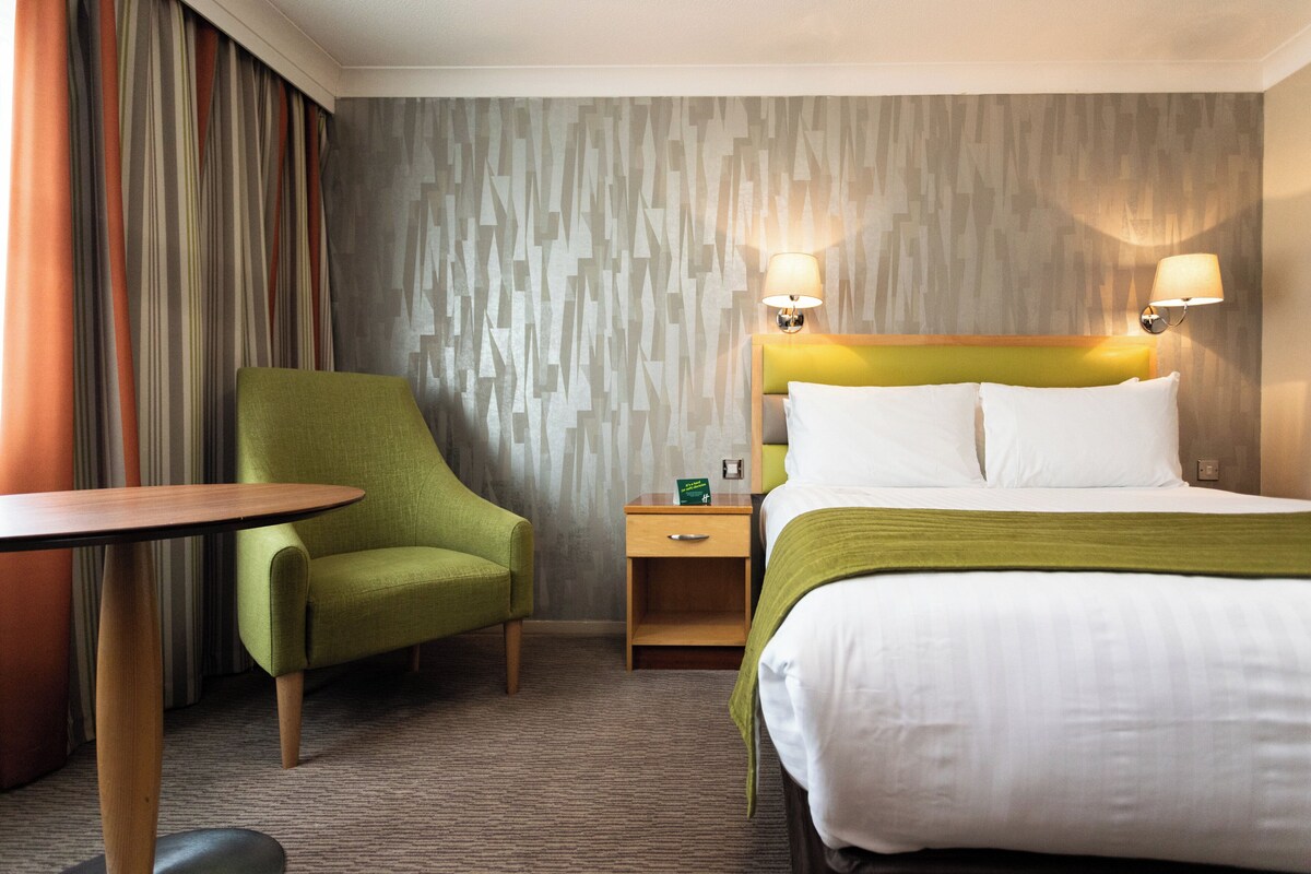 Une chambre d’hôtel avec un lit, un fauteuil vert, une table de chevet en bois et une table.