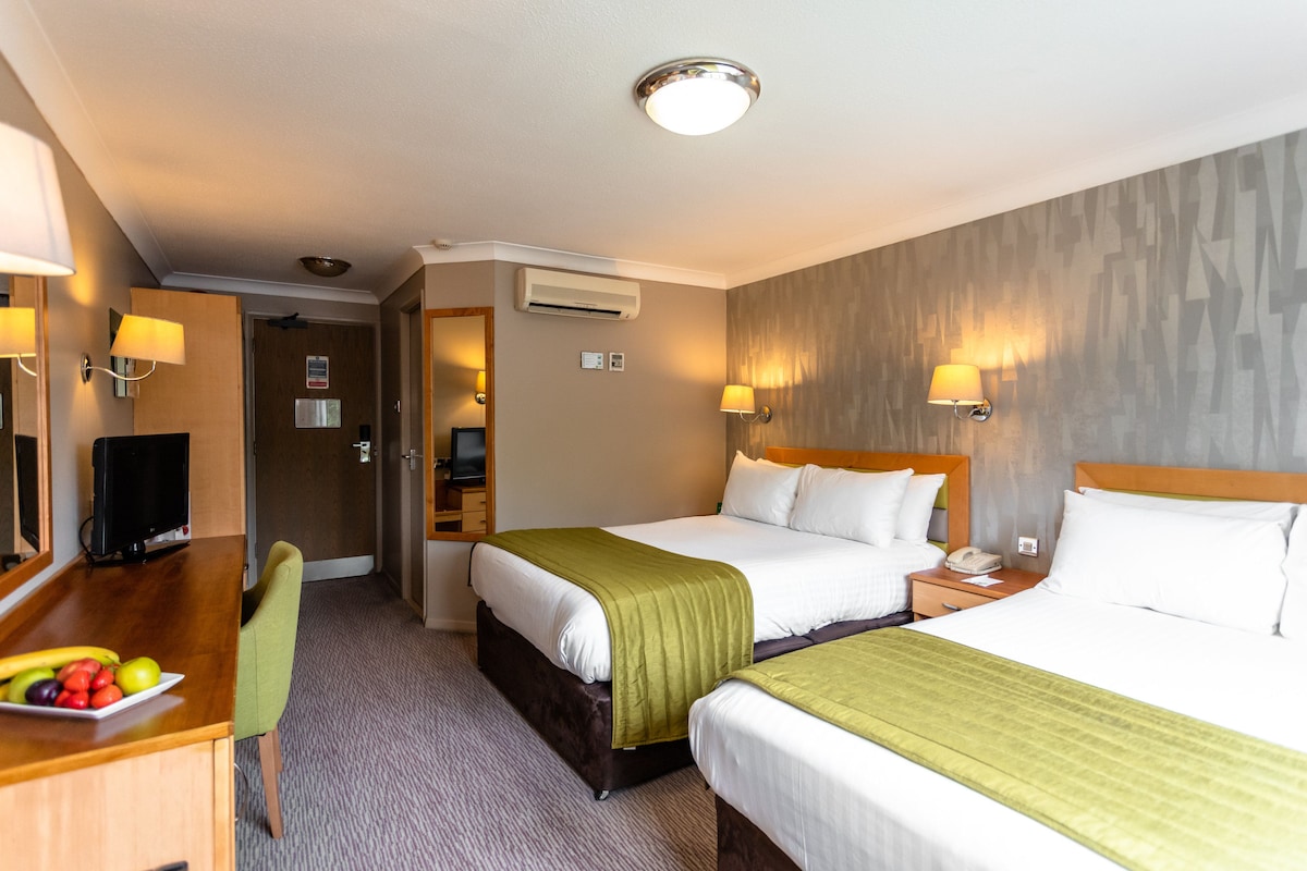 A hotel room with two beds, a desk with a television, a chair, and a fruit bowl.