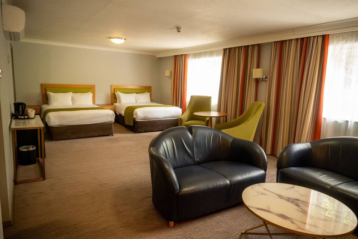 Habitación de hotel con dos camas, un sillón verde, un sofá de cuero negro y una pequeña mesa redonda.