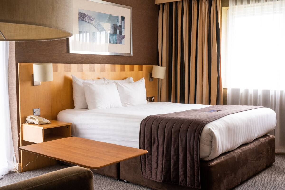 Una habitación de hotel con una cama grande, una mesita de noche, una lámpara, un teléfono y una ventana con cortinas.